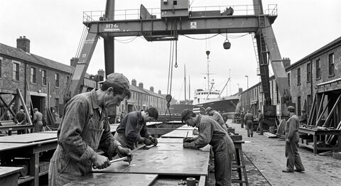 Shipyard workers in overalls riveting steel plates under a tall gantry crane in an old Clyde yard.