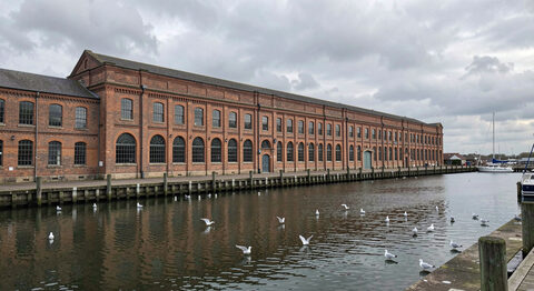A long Victorian brick warehouse with arched windows beside a dock basin under a cloudy sky.